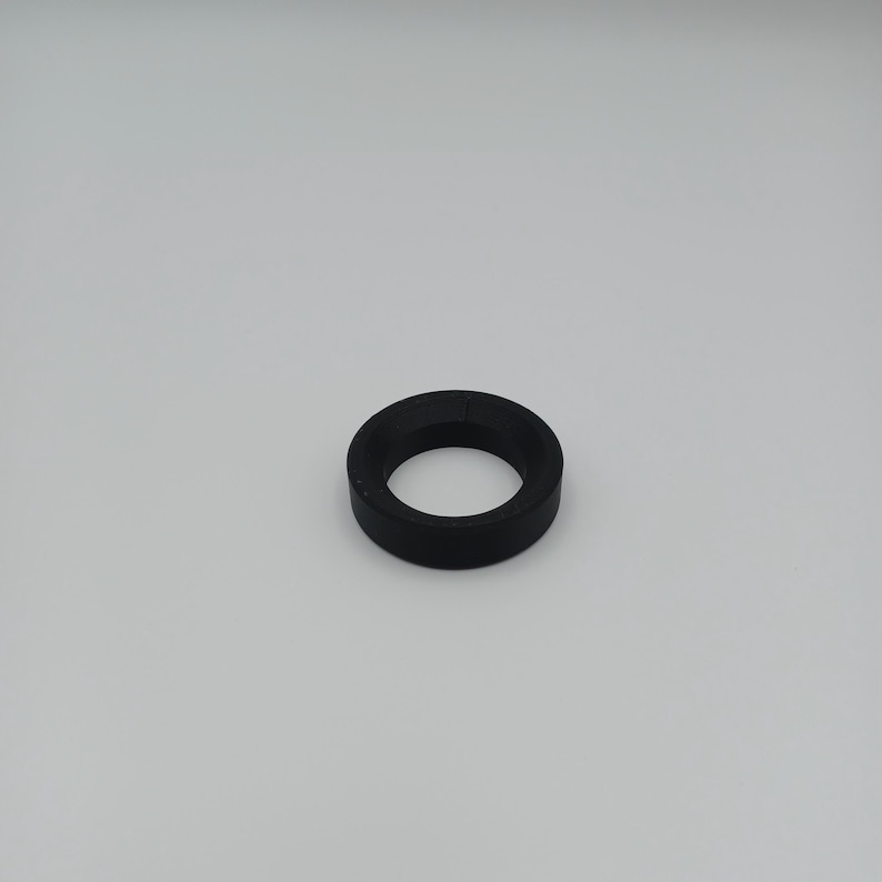 May include: A black, circular rubber gasket with a hole in the center. The gasket is made of a solid, matte material and is designed to create a seal. The object is set against a plain white background.