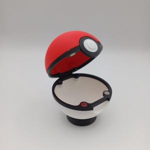 May include: A red and white Pok&eacute; Ball replica, open to reveal a white interior. The top half is red, the bottom half is white, and a black band separates the two. A black base supports the ball.