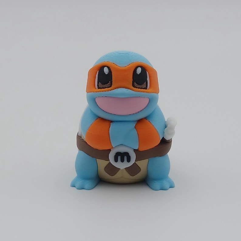 May include: A blue and orange 3D printed figurine of a turtle wearing a bandana with the letter "M" on its chest.