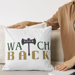 Puede incluir: Cojín blanco con el texto "WATCH YOUR BACK" en letras verdes y doradas. Un hacha negra con la palabra "YOUR" en ella está en el centro de la almohada.