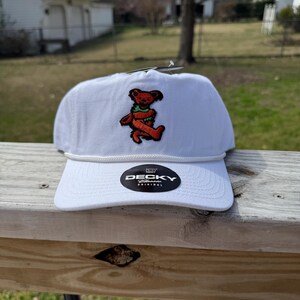 May include: A white baseball cap with a red bear graphic on the front. The cap has a curved brim, a white rope detail, and a black and white "DECKY" label. The hat is made of a durable material and is suitable for outdoor wear.