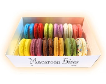 May include: A box of colorful macarons, including blue, red, yellow, green, purple, and white. The macarons are arranged in two rows inside a white box with the text "Macaroon Bites". Each macaron is approximately 2 inches in diameter.