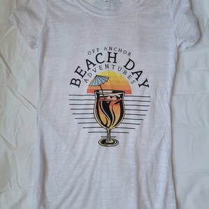 May include: White t-shirt with a graphic of a cocktail drink with an umbrella and the text "Off Anchor Beach Day Adventures".