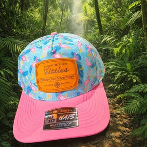 May include: A blue and pink baseball cap featuring a flamingo design and the text "SHOW ME THEM Titties". The cap has a pink peak and a patch that reads "Off Anchor Adventures". The hat is waterproof and adjustable.