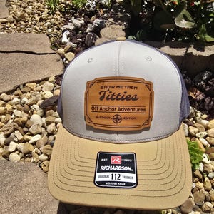 May include: A grey and brown trucker hat with a brown leather patch that says "Show Me Them Titties Off Anchor Adventures Outdoor Edition". The hat also has a brown patch that says "Est. 1970 Richardson Original 112 Trucker Adjustable".