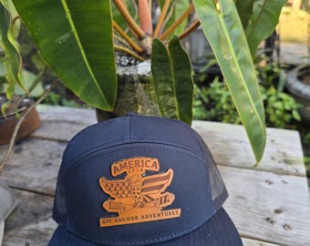 May include: A navy blue snapback hat with a flat bill and a mesh back. A brown leather patch on the front features an eagle graphic, the text "AMERICA 1776", and "OFF ANCHOR ADVENTURES". A black and white sticker on the bill reads "RICHARDSON".