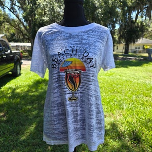 May include: White t-shirt with a distressed, faded look. The shirt features a graphic of a cocktail glass with a sunset and palm tree behind it. The text "Off Anchor Beach Day Adventures" is printed around the graphic.