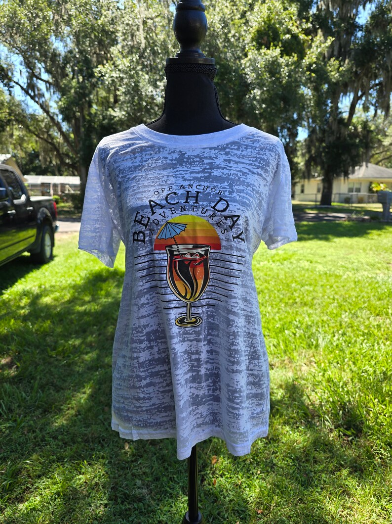 May include: White t-shirt with a distressed, faded look. The shirt features a graphic of a cocktail glass with a sunset and palm tree behind it. The text "Off Anchor Beach Day Adventures" is printed around the graphic.