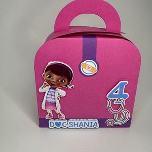 Może przedstawiać: Różowe pudełko na upominki z uchwytem, z wizerunkiem kreskówki Doc McStuffins. Pudełko ma fioletowy pasek pośrodku, naklejkę z plastrem i cyfrę 4 ze stetoskopem. Nazwa "DOC SHANIA" jest wydrukowana na dole.