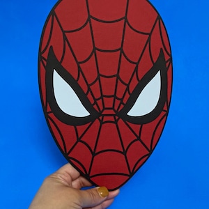 Puede incluir: Una m&aacute;scara roja de Spider-Man con un dise&ntilde;o de telara&ntilde;a negra y formas de ojos blancos. La m&aacute;scara se sostiene sobre un fondo azul. El objeto es de tem&aacute;tica de superh&eacute;roes.