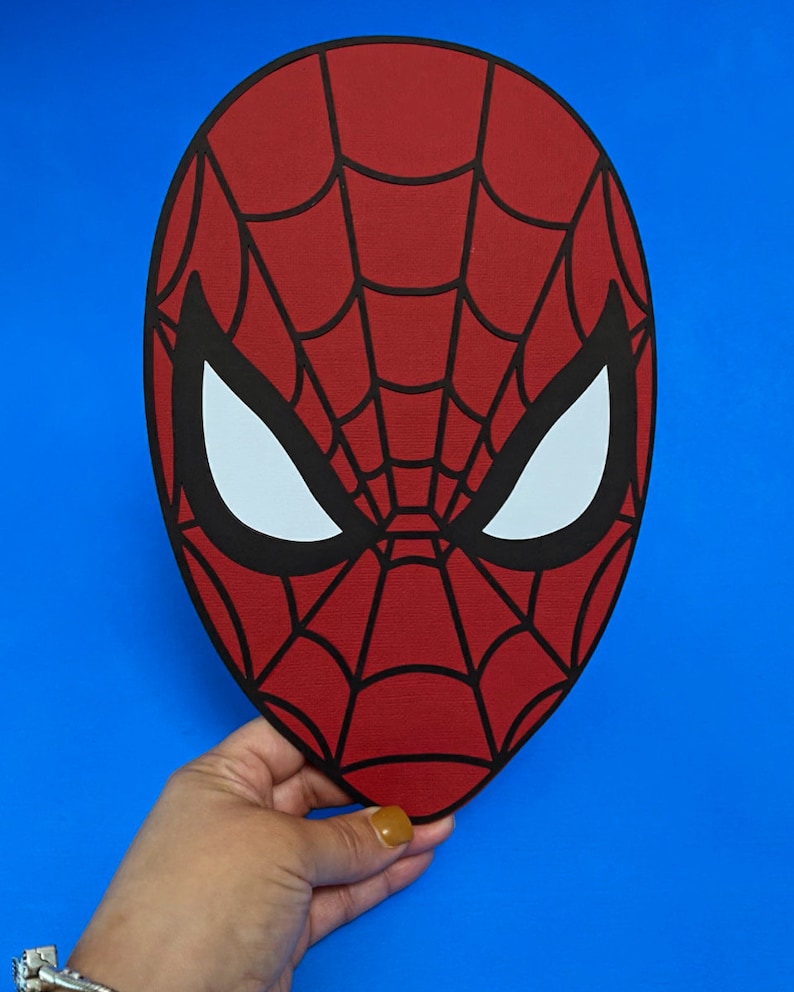Puede incluir: Una m&aacute;scara roja de Spider-Man con un dise&ntilde;o de telara&ntilde;a negra y formas de ojos blancos. La m&aacute;scara se sostiene sobre un fondo azul. El objeto es de tem&aacute;tica de superh&eacute;roes.
