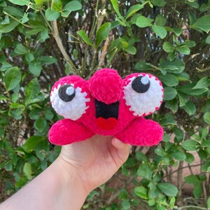 Peut inclure: Une cr&eacute;ature rose en peluche faite &agrave; la main, avec de grands yeux bord&eacute;s de blanc et une bouche noire. Le jouet est tenu dans la main d'une personne sur fond de feuillage vert. La cr&eacute;ature a une texture floue.