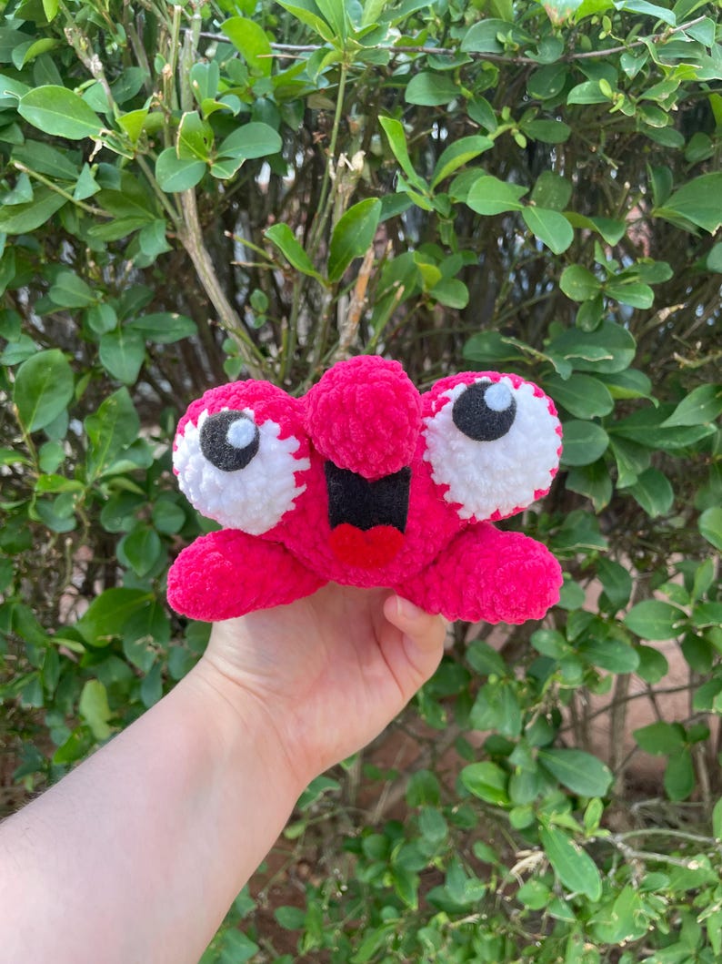 Peut inclure: Une cr&eacute;ature rose en peluche faite &agrave; la main, avec de grands yeux bord&eacute;s de blanc et une bouche noire. Le jouet est tenu dans la main d'une personne sur fond de feuillage vert. La cr&eacute;ature a une texture floue.