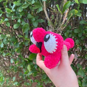 Peut inclure: Une cr&eacute;ature rose vif faite &agrave; la main, crochet&eacute;e, avec des yeux blancs et noirs et une bouche rouge. La peluche est tenue dans une main sur fond de feuillage vert. Le jouet a un design fantaisiste et cartoon.