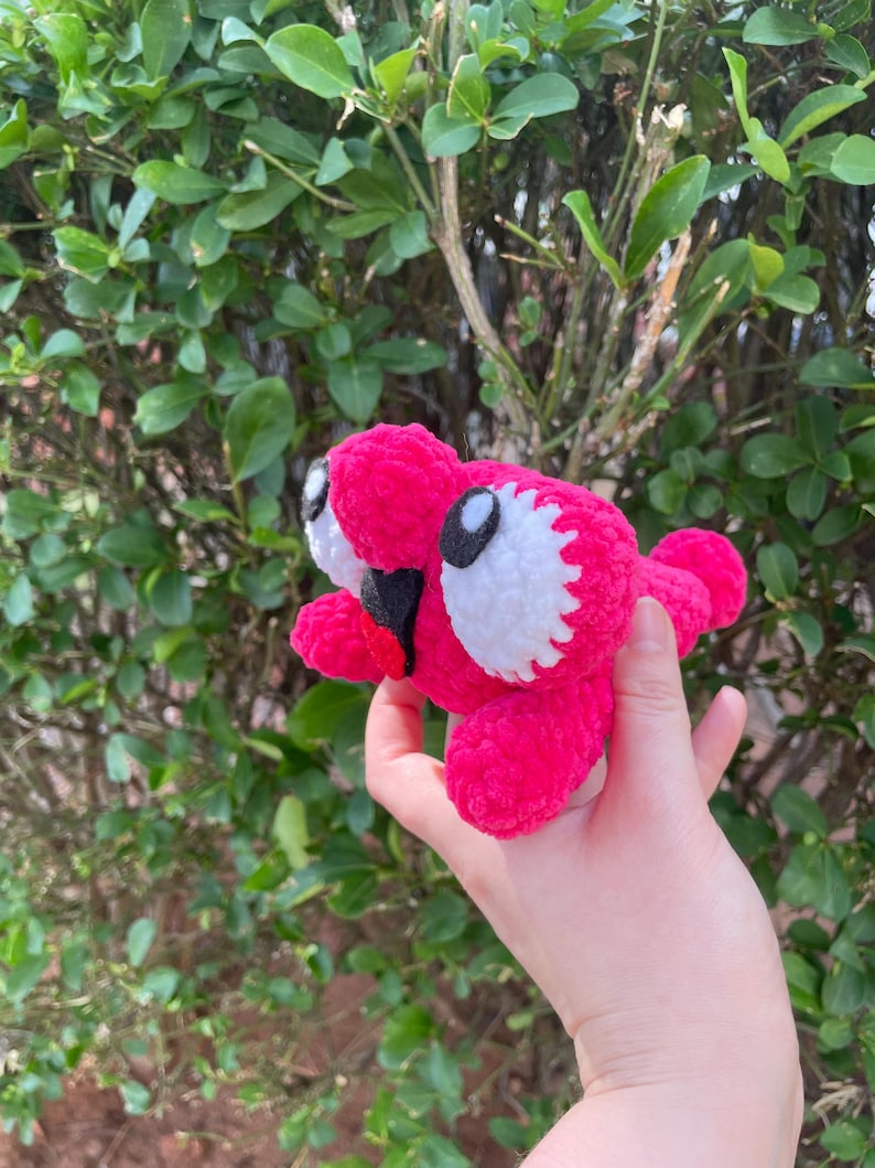 Peut inclure: Une cr&eacute;ature rose vif faite &agrave; la main, crochet&eacute;e, avec des yeux blancs et noirs et une bouche rouge. La peluche est tenue dans une main sur fond de feuillage vert. Le jouet a un design fantaisiste et cartoon.