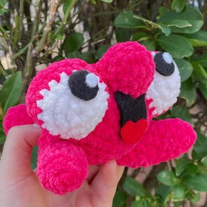 Peut inclure: Un jouet en peluche fait à la main, rose vif, avec de grands yeux blancs et noirs et une bouche en forme de cœur rouge. Le jouet est tenu devant un fond de feuillage vert, mettant en valeur ses couleurs vives et sa texture douce.