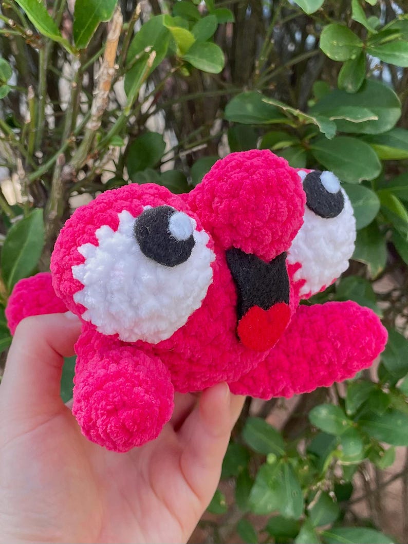 Peut inclure: Un jouet en peluche fait &agrave; la main, rose vif, avec de grands yeux blancs et noirs et une bouche en forme de c&oelig;ur rouge. Le jouet est tenu devant un fond de feuillage vert, mettant en valeur ses couleurs vives et sa texture douce.