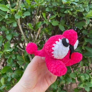Peut inclure: Une cr&eacute;ature au crochet rose vif faite &agrave; la main avec des accents blancs et noirs. Le jouet en peluche a de grands yeux ronds et un petit bec. La cr&eacute;ature est tenue devant un fond de feuillage vert.