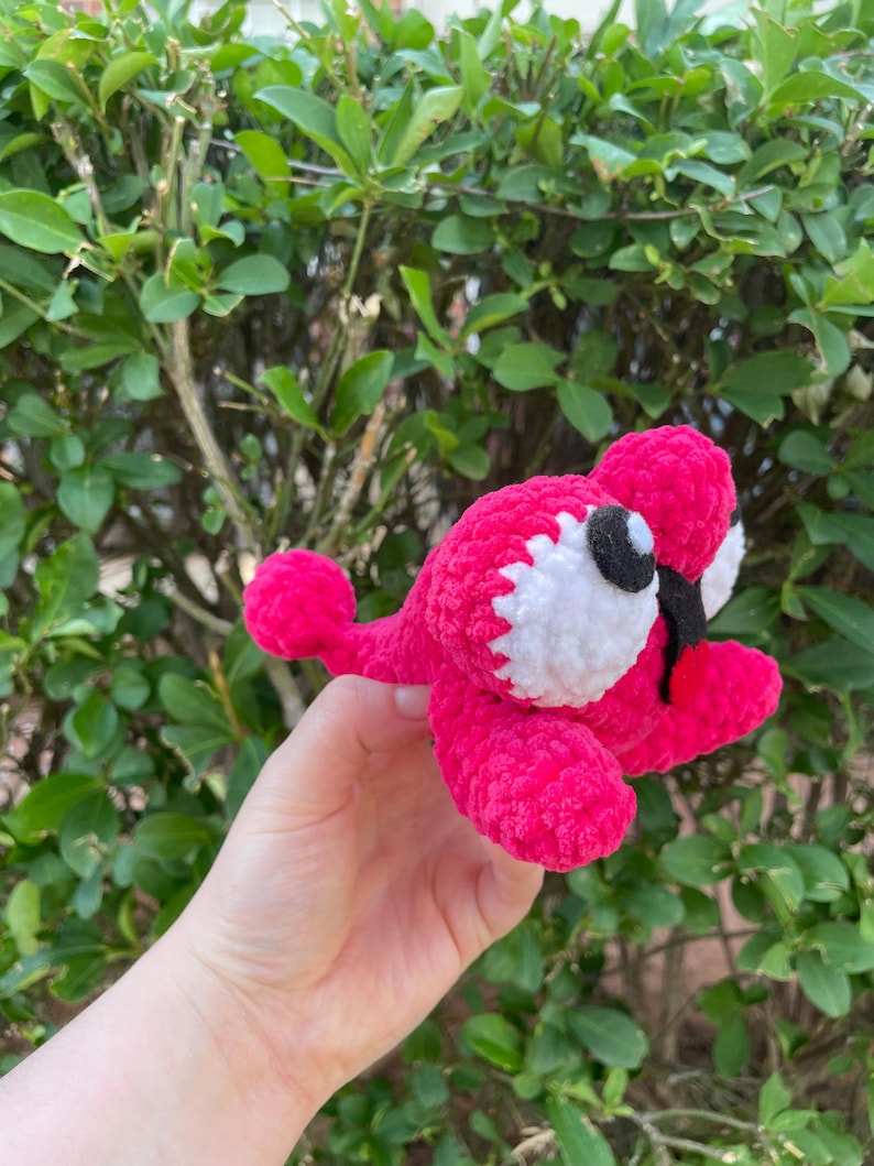 Peut inclure: Une cr&eacute;ature au crochet rose vif faite &agrave; la main avec des accents blancs et noirs. Le jouet en peluche a de grands yeux ronds et un petit bec. La cr&eacute;ature est tenue devant un fond de feuillage vert.