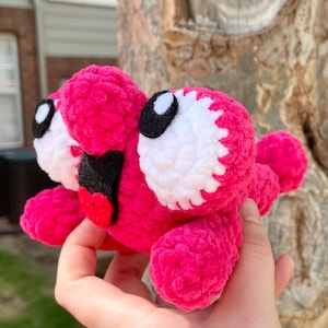 Peut inclure: Une cr&eacute;ature rose en peluche faite &agrave; la main avec de grands yeux blancs et noirs, une bouche noire et une langue rouge. Le jouet doux au crochet a de multiples appendices et un design fantaisiste, id&eacute;al pour une collection de jouets pour enfants.