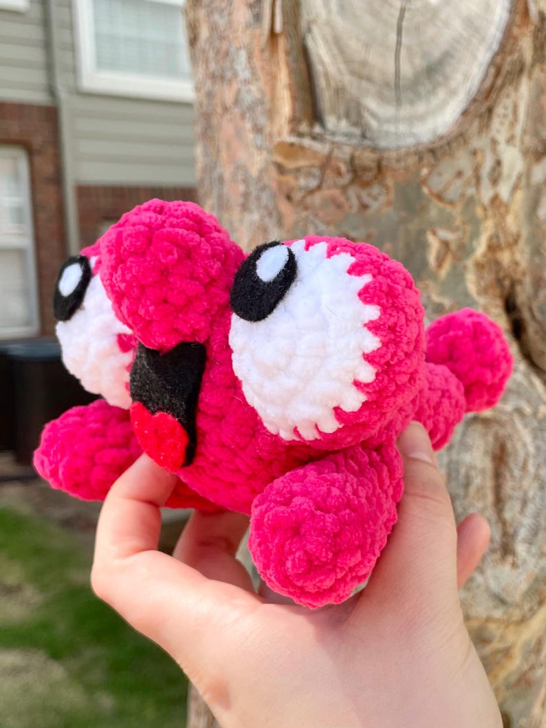 Peut inclure: Une cr&eacute;ature rose en peluche faite &agrave; la main avec de grands yeux blancs et noirs, une bouche noire et une langue rouge. Le jouet doux au crochet a de multiples appendices et un design fantaisiste, id&eacute;al pour une collection de jouets pour enfants.