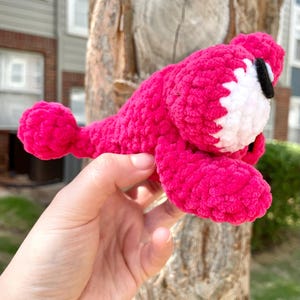 Peut inclure: Un animal rose vif fait &agrave; la main, crochet&eacute;, avec des accents blancs et noirs. Le jouet en peluche a un corps arrondi, une petite queue et un visage blanc avec des yeux noirs. Le jouet est tenu dans une main.