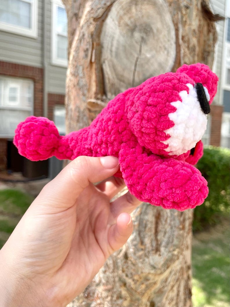 Peut inclure: Un animal rose vif fait &agrave; la main, crochet&eacute;, avec des accents blancs et noirs. Le jouet en peluche a un corps arrondi, une petite queue et un visage blanc avec des yeux noirs. Le jouet est tenu dans une main.