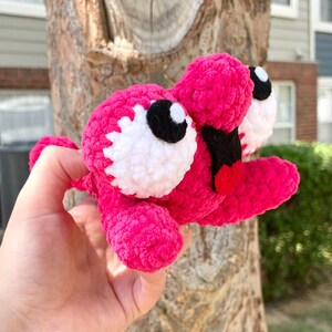 Peut inclure: Une cr&eacute;ature au crochet rose vif faite &agrave; la main, avec de grands yeux blancs et noirs, une bouche noire et une langue rouge. Le jouet en peluche est tenu devant un tronc d'arbre. Le corps de la cr&eacute;ature est fait de fil doux et textur&eacute;.