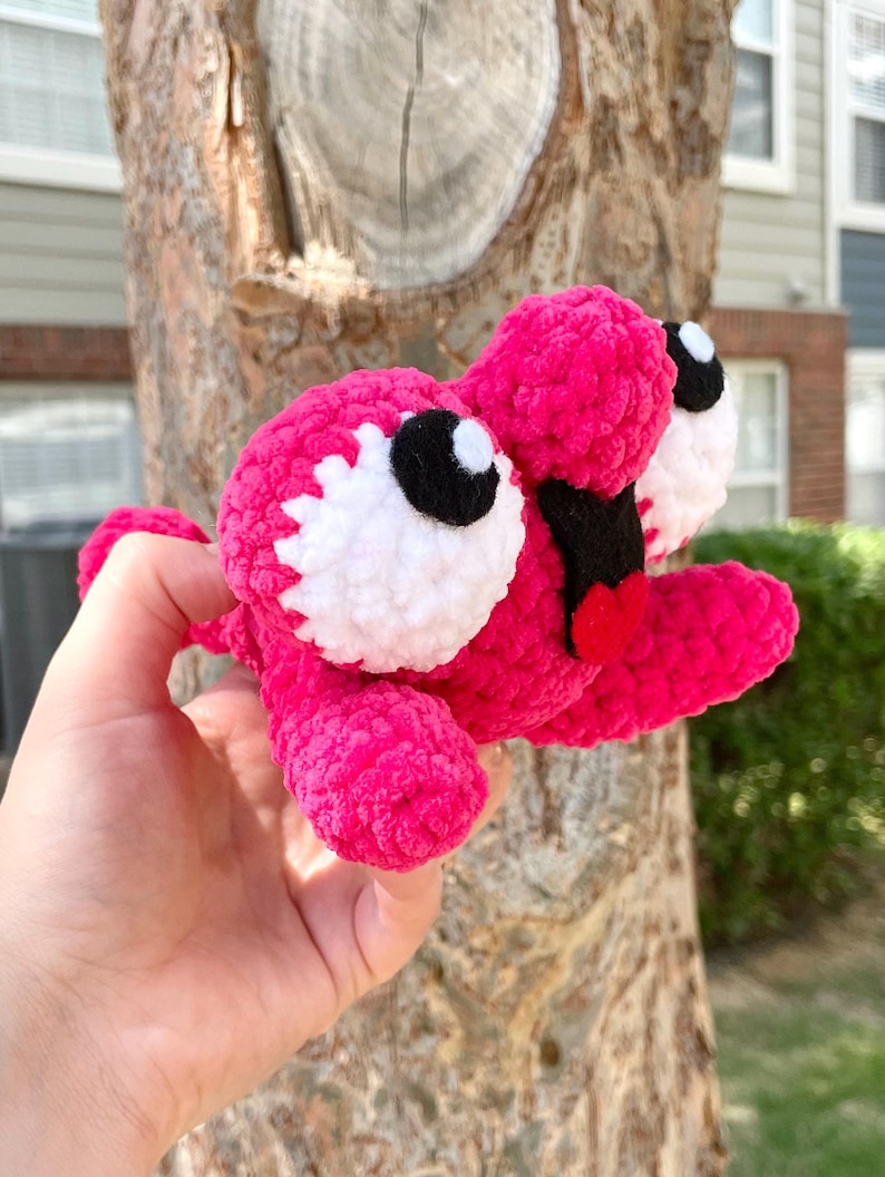 Peut inclure: Une cr&eacute;ature au crochet rose vif faite &agrave; la main, avec de grands yeux blancs et noirs, une bouche noire et une langue rouge. Le jouet en peluche est tenu devant un tronc d'arbre. Le corps de la cr&eacute;ature est fait de fil doux et textur&eacute;.