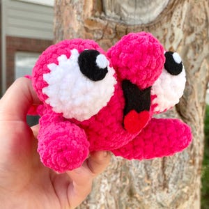 Peut inclure: Une cr&eacute;ature rose en peluche faite &agrave; la main, avec de grands yeux blancs et noirs, une bouche noire et une langue rouge. Le jouet doux et textur&eacute; est tenu dans une main, mettant en valeur ses couleurs vives et son design fantaisiste. Le corps de la cr&eacute;ature est rose vif.