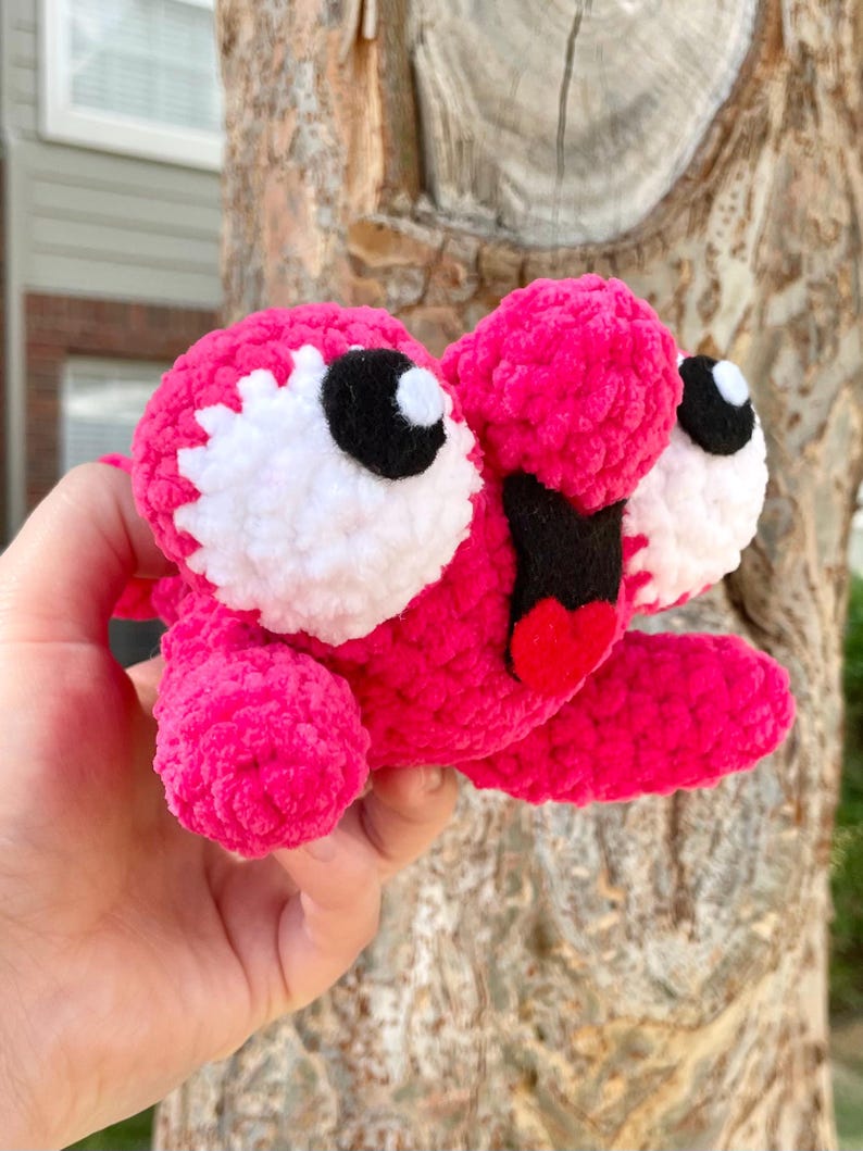Peut inclure: Une cr&eacute;ature rose en peluche faite &agrave; la main, avec de grands yeux blancs et noirs, une bouche noire et une langue rouge. Le jouet doux et textur&eacute; est tenu dans une main, mettant en valeur ses couleurs vives et son design fantaisiste. Le corps de la cr&eacute;ature est rose vif.