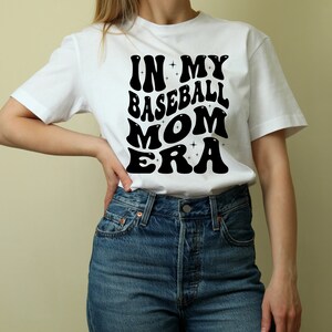 May include: White t-shirt with black text that reads "In My Baseball Mom Era" in a retro font.