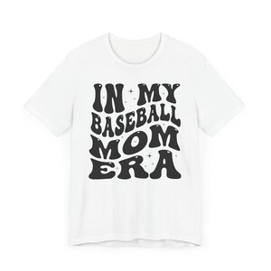May include: White t-shirt with black text that reads "In My Baseball Mom Era" in a retro font.