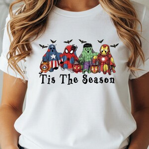 Puede incluir: Camiseta blanca con un dise&ntilde;o de superh&eacute;roes disfrazados de fantasmas, sosteniendo linternas, con murci&eacute;lagos encima. El texto "Tis The Season" est&aacute; impreso debajo. La camiseta tiene cuello redondo y mangas enrolladas.