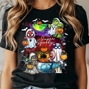 May include: Black t-shirt featuring a colorful Halloween graphic with superheroes dressed as ghosts and holding jack-o'-lanterns. The text "Super Spooky" is written in a script font. The design includes bats, spiderwebs, and a spooky forest scene.