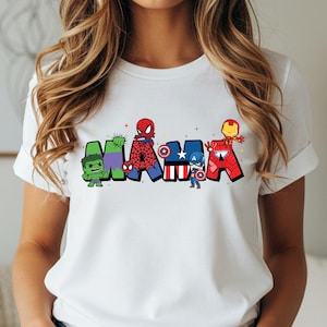 May include: White t-shirt with the word 'MAMA' in colorful superhero-themed letters. The letters are designed with characters like Hulk, Spider-Man, Captain America, and Iron Man. The shirt is a casual wear item, perfect for fans of Marvel comics and superheroes.