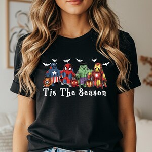 Puede incluir: Camiseta negra con un dise&ntilde;o de personajes de superh&eacute;roes, incluyendo al Capit&aacute;n Am&eacute;rica, Spider-Man, Hulk e Iron Man, disfrazados de fantasmas. La frase "Tis The Season" est&aacute; impresa debajo, con siluetas de murci&eacute;lagos arriba.