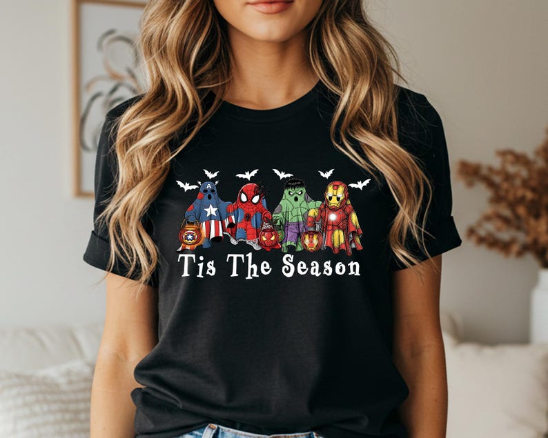 Puede incluir: Camiseta negra con un dise&ntilde;o de personajes de superh&eacute;roes, incluyendo al Capit&aacute;n Am&eacute;rica, Spider-Man, Hulk e Iron Man, disfrazados de fantasmas. La frase "Tis The Season" est&aacute; impresa debajo, con siluetas de murci&eacute;lagos arriba.