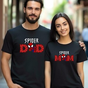 May include: Black t-shirts with Spider-Man designs. One shirt says "SPIDER DAD" with a Spider-Man graphic, and the other says "SPIDER MOM" with a Spider-Man graphic. The text is in white and red.