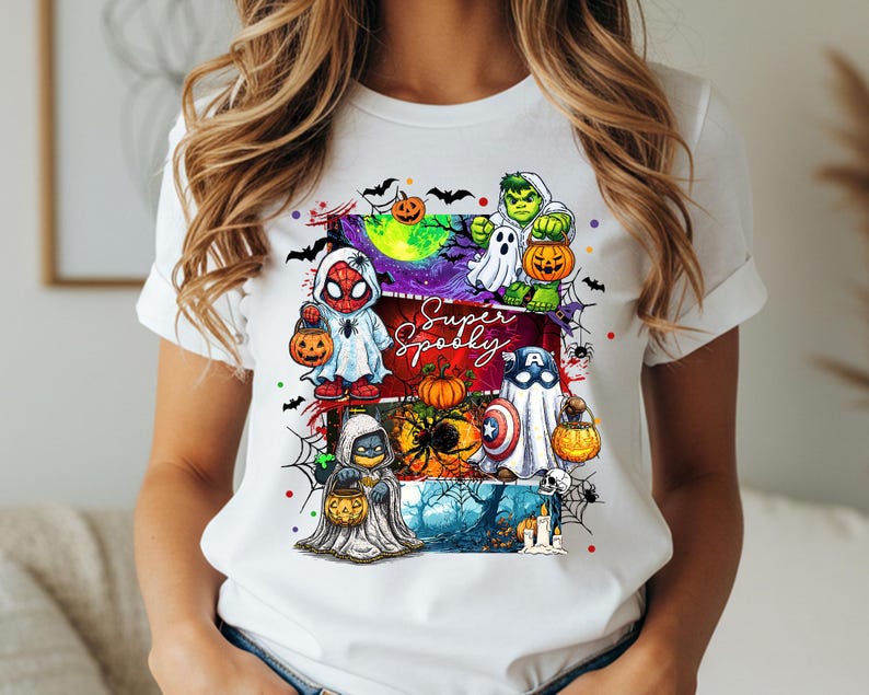 May include: White t-shirt featuring a colorful Halloween-themed graphic. The design includes cartoon characters dressed as superheroes, pumpkins, ghosts, and the text "Super Spooky". Bats, spiderwebs, and a spooky night scene complete the design.