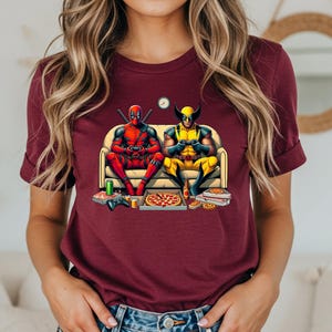 May include: Burgundy t-shirt featuring a graphic of Deadpool and Wolverine on a couch, playing video games. Pizza boxes and a pizza are on the floor. A small clock is on the wall. The characters are in their superhero costumes.