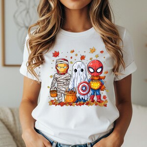 May include: White t-shirt featuring a Halloween-themed graphic. The design includes cartoon versions of Iron Man as a mummy, a ghost, and Spider-Man, all holding pumpkin buckets. Autumn leaves surround the characters.