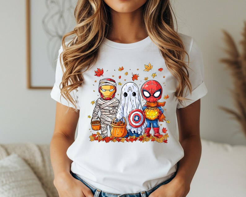 May include: White t-shirt featuring a Halloween-themed graphic. The design includes cartoon versions of Iron Man as a mummy, a ghost, and Spider-Man, all holding pumpkin buckets. Autumn leaves surround the characters.