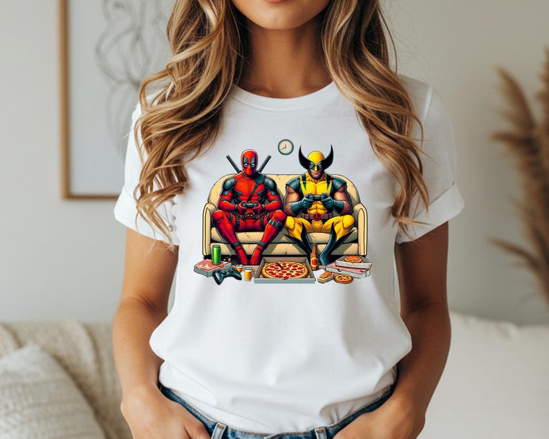 May include: White t-shirt featuring a colorful graphic of Deadpool and Wolverine relaxing on a couch, playing video games, and eating pizza. The characters are in their superhero costumes, with Deadpool in red and Wolverine in yellow and blue. Pizza boxes, controllers, and a clock are also visible.