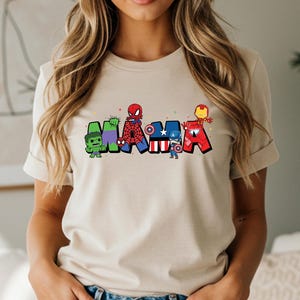 Puede incluir: Camiseta beige con la palabra "MAMA" en letras coloridas con tem&aacute;tica de superh&eacute;roes. Cada letra presenta un personaje diferente: Hulk, Spider-Man, Capit&aacute;n Am&eacute;rica e Iron Man. El dise&ntilde;o incluye estrellas y est&aacute; impreso en una camiseta de manga corta.