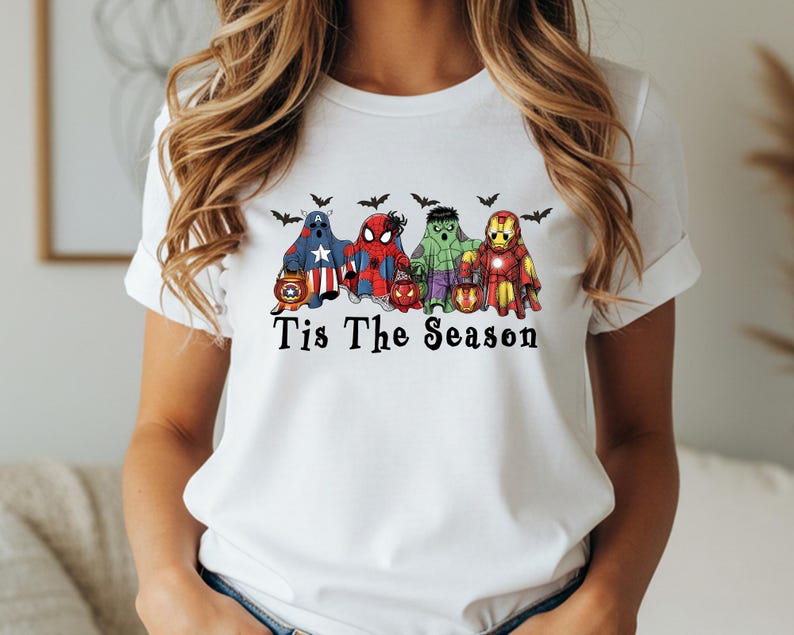 Puede incluir: Camiseta blanca con un dise&ntilde;o de superh&eacute;roes disfrazados de fantasmas, sosteniendo linternas, con murci&eacute;lagos encima. El texto "Tis The Season" est&aacute; impreso debajo. La camiseta tiene cuello redondo y mangas enrolladas.
