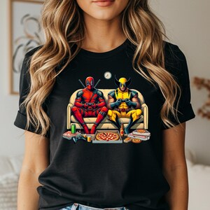 May include: Black t-shirt featuring a colorful graphic of Deadpool and Wolverine playing video games on a couch. Pizza boxes, controllers, and soda cans are on the floor. A clock is on the wall.