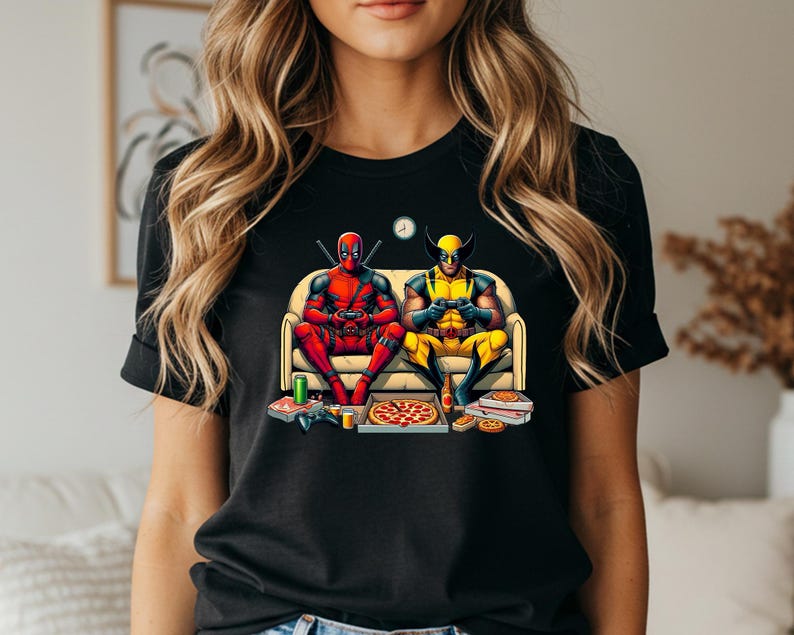 May include: Black t-shirt featuring a colorful graphic of Deadpool and Wolverine playing video games on a couch. Pizza boxes, controllers, and soda cans are on the floor. A clock is on the wall.