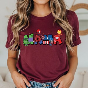 Puede incluir: Una camiseta granate con la palabra "MAMA" en letras coloridas de estilo caricatura, cada letra dise&ntilde;ada con un tema de superh&eacute;roe diferente. La camiseta la lleva una persona con pelo largo y rubio ondulado.