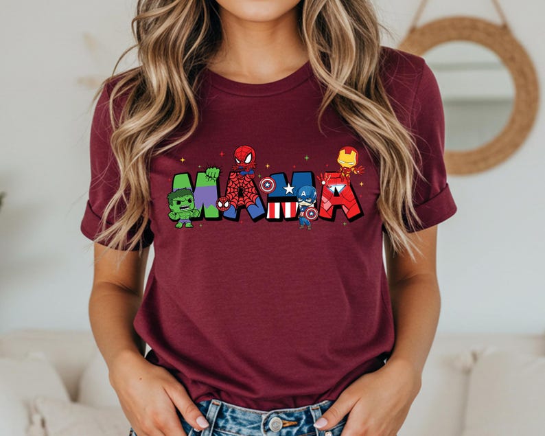 Puede incluir: Una camiseta granate con la palabra "MAMA" en letras coloridas de estilo caricatura, cada letra dise&ntilde;ada con un tema de superh&eacute;roe diferente. La camiseta la lleva una persona con pelo largo y rubio ondulado.